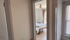 Un pasillo que conduce a un dormitorio con una cama, una lámpara de noche y una ventana con persianas.