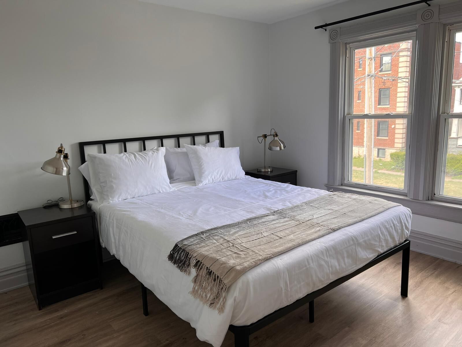 A bedroom with a bed, two bedside tables, a window with curtains, and a view of buildings outside.