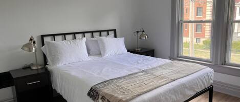 A bedroom with a bed, two bedside tables, a window with curtains, and a view of buildings outside.