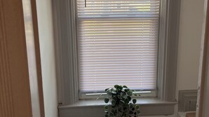 Un ventanal de esquina con persianas blancas, una planta pequeña colgando del alféizar y un suelo de madera.