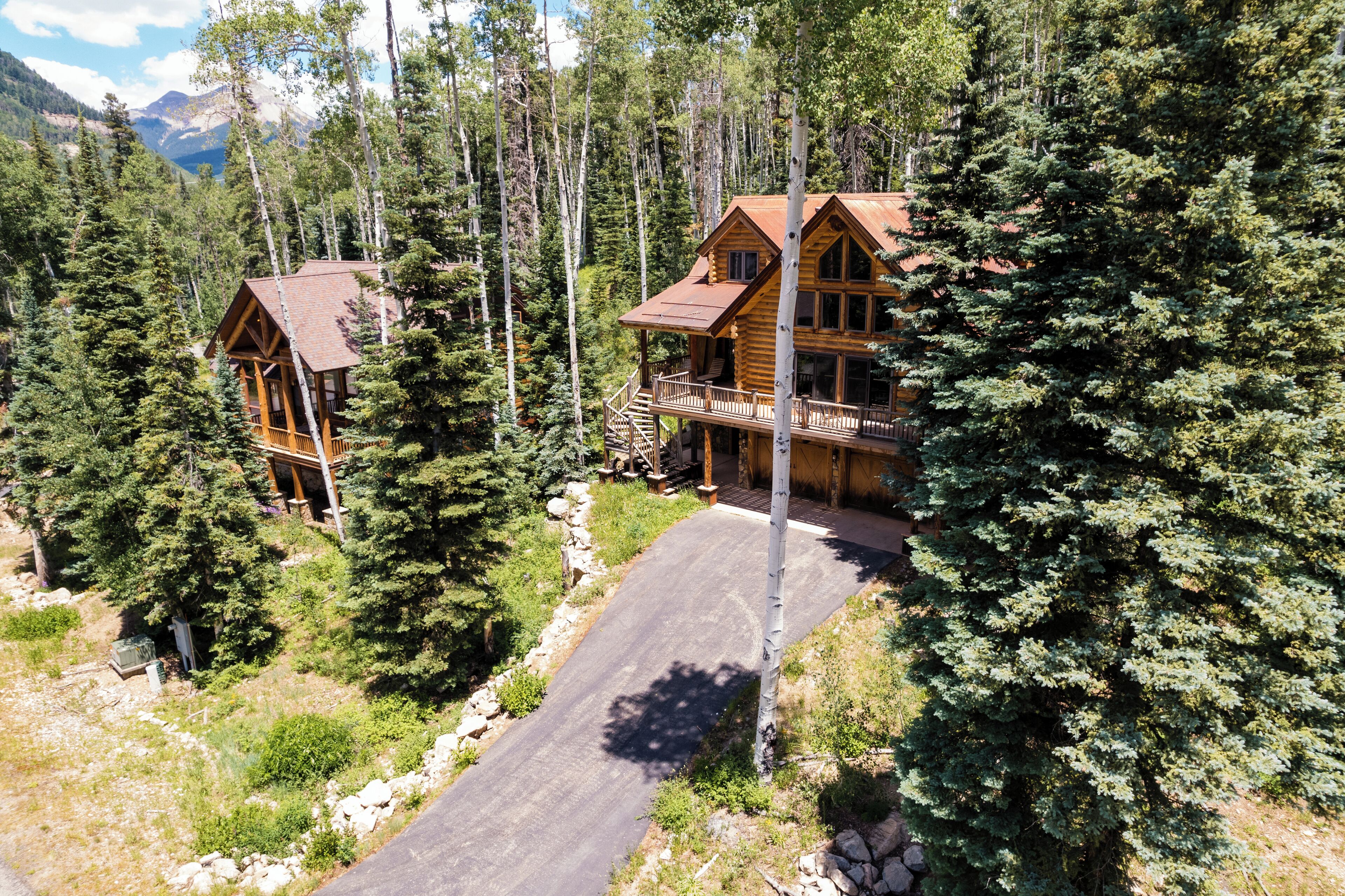 A wooden cabin with a balcony, surrounded by dense forest and mountains in the background.