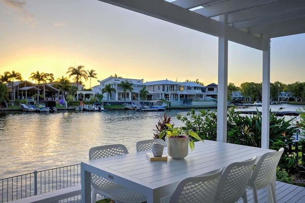 Un patio frente al río con una mesa y sillas, con vista a un río tranquilo donde se ven barcos y casas al fondo.