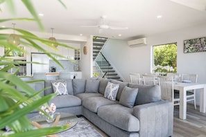 A modern living room with a grey sectional sofa, a dining table with chairs, a staircase, and a kitchen in the background.