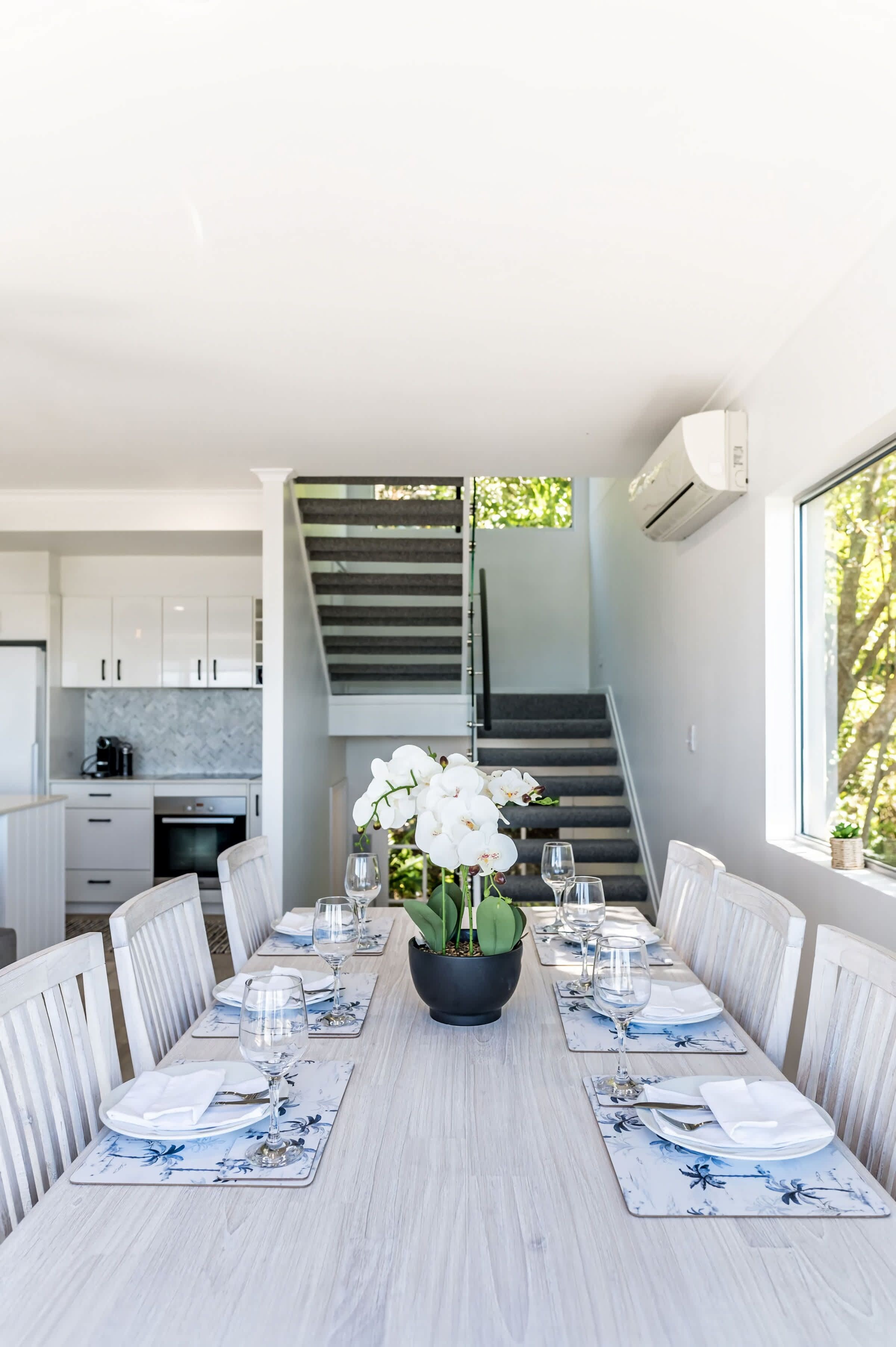 Uma sala de jantar moderna com uma mesa comprida posta para refeição, uma escada e uma cozinha ao fundo.
