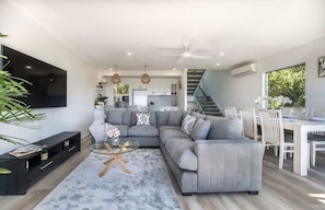 Uma sala de estar moderna com um sofá modular cinza, uma mesa de centro de vidro e uma área de jantar com mesa e cadeiras brancas.
