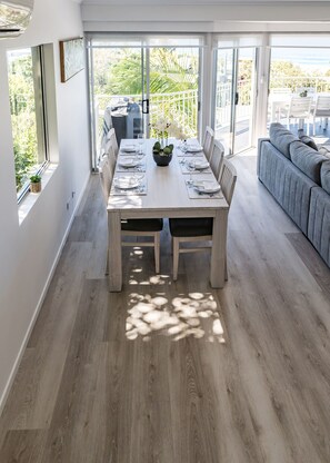 Uma sala de jantar com mesa de madeira posta para seis, grandes janelas e um sofá cinza ao fundo.