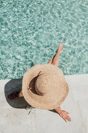 Une personne se détend au bord d’une piscine, portant un chapeau de paille, l’eau est d’un bleu limpide et le bord de la piscine est en béton.