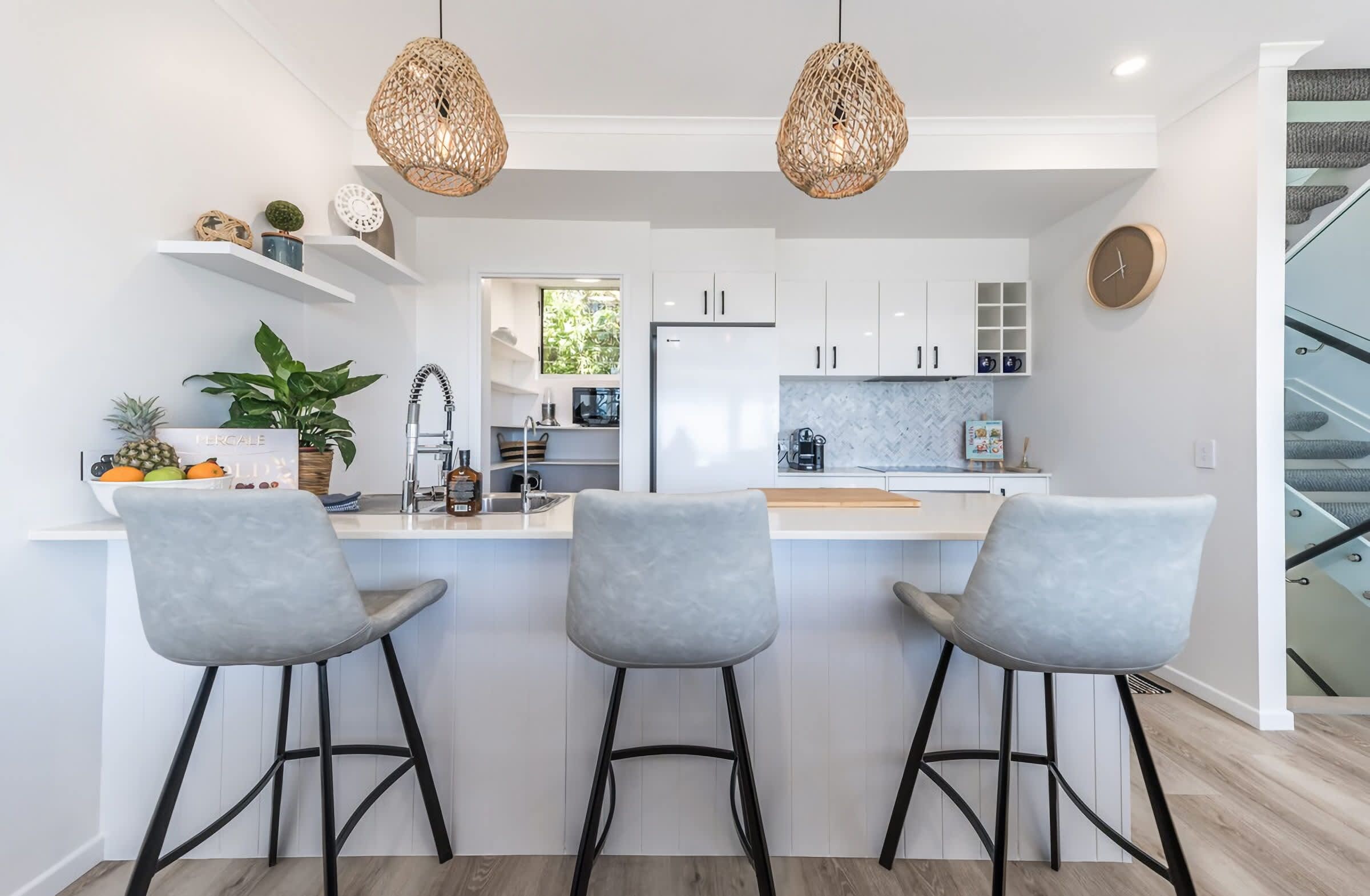 A modern kitchen with white cabinets, a central island with bar stools, a hanging light fixture, and a clock on the wall.