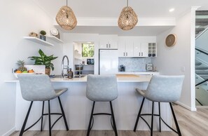 Une cuisine moderne avec des meubles blancs, un îlot central doté de tabourets, un luminaire suspendu et une horloge au mur.