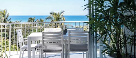 Un balcon avec des chaises blanches et une table, d’où l’on peut admirer une mer bleue et limpide ainsi que des palmiers.