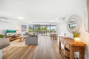 A modern living room with a sofa, TV, and dining area, large windows, and a wooden floor.