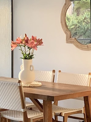 Une table à manger avec un vase blanc rempli de fleurs roses, des chaises en bois et un miroir rond accroché au mur.