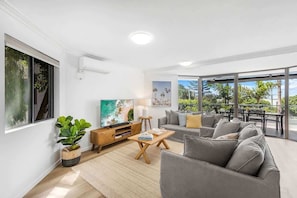 Un salon moderne avec un canapé, une table basse, un téléviseur et une grande fenêtre donnant sur un balcon.