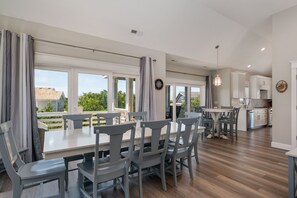 A spacious dining area with a large table and chairs, a kitchen with white cabinets, and a view through large windows. - Benders Beach Bungalow: New Construction, Golf Views, Elevator, Community Pool and Tennis Courts (Nags Head)