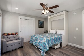 A bedroom with a bed, a gray sofa, a ceiling fan, and a door. - Benders Beach Bungalow: New Construction, Golf Views, Elevator, Community Pool and Tennis Courts (Nags Head)