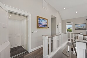 A hallway with a door, a painting, and a staircase with a railing. - Benders Beach Bungalow: New Construction, Golf Views, Elevator, Community Pool and Tennis Courts (Nags Head)