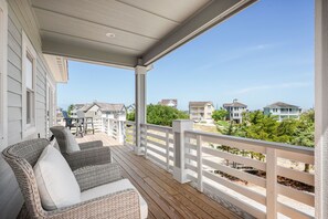 A covered outdoor deck with wicker furniture and a white railing, overlooking a residential area with houses and greenery. - Benders Beach Bungalow: New Construction, Golf Views, Elevator, Community Pool and Tennis Courts (Nags Head)