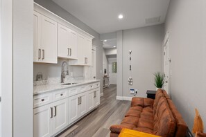 A modern kitchen with white cabinets and a marble countertop, leading to a living area with a brown leather sofa. - Benders Beach Bungalow: New Construction, Golf Views, Elevator, Community Pool and Tennis Courts (Nags Head)