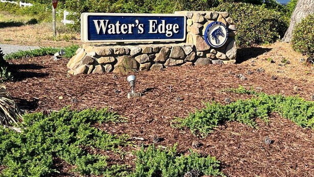 A sign reading "Water's Edge" with a stone wall and a decorative emblem. - Benders Beach Bungalow: New Construction, Golf Views, Elevator, Community Pool and Tennis Courts (Nags Head)