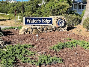 A sign reading "Water's Edge" with a stone wall and a decorative emblem. - Benders Beach Bungalow: New Construction, Golf Views, Elevator, Community Pool and Tennis Courts (Nags Head)
