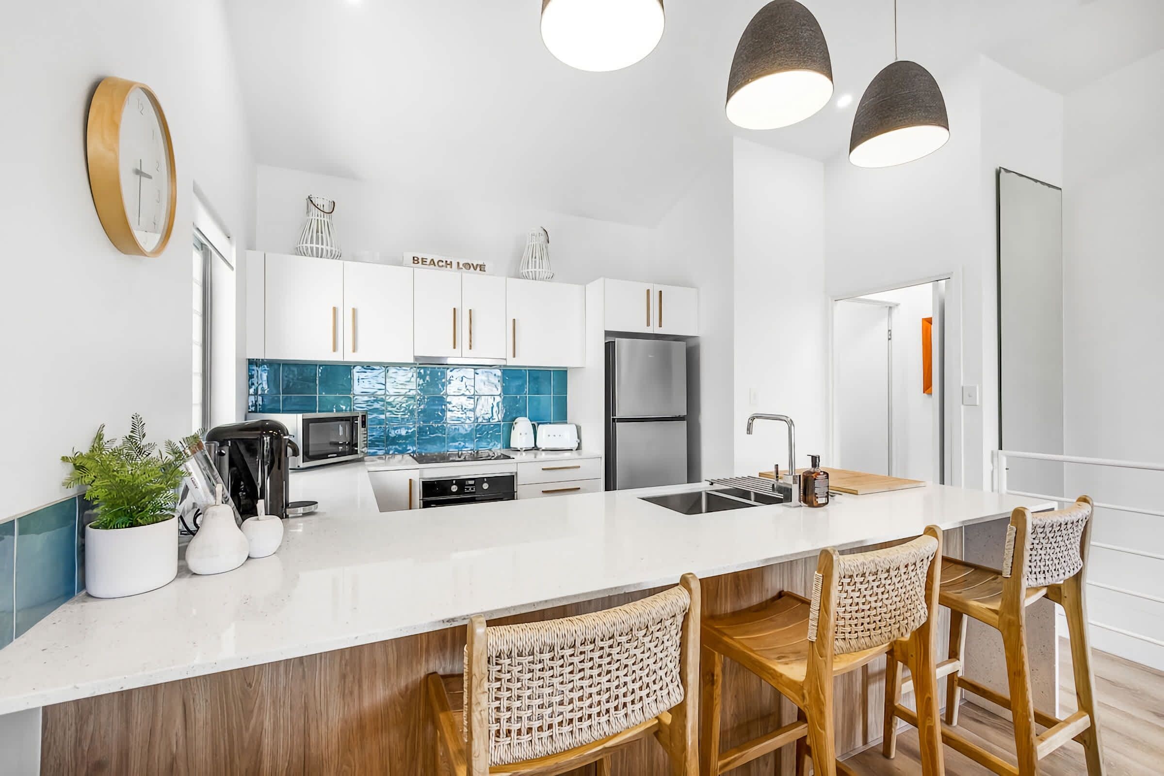 Une cuisine moderne avec des meubles blancs, un îlot central doté de tabourets de bar, des appareils en acier inoxydable et un éclairage suspendu.
