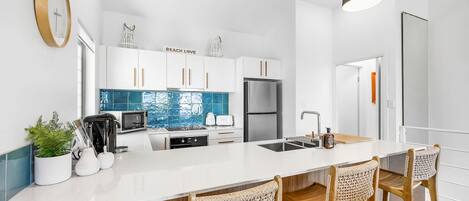 Une cuisine moderne avec des meubles blancs, un îlot central doté de tabourets de bar, des appareils en acier inoxydable et un éclairage suspendu.
