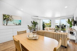 A modern living room with a dining area, featuring a wooden table, wicker chairs, a sofa, and a large window with a view of greenery.