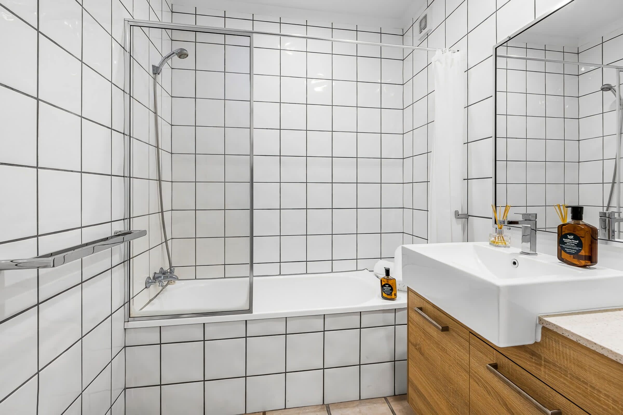 Un baño moderno con paredes de azulejos blancos, una bañera, zona de ducha y tocador con espejo.