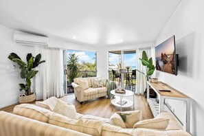A living room with a sofa, coffee table, TV, and large windows with a view. - 3 Andari Sunshine Beach Modern Apt 1 Min Beach (Noosa)