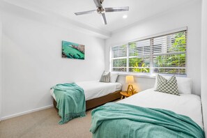 A bedroom with two beds, a ceiling fan, a window with blinds, and a painting on the wall.