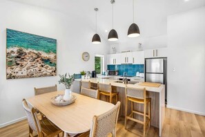 A modern kitchen with white cabinets, a stainless steel refrigerator, and a dining area with a wooden table and wicker chairs. - 3 Andari Sunshine Beach Modern Apt 1 Min Beach (Noosa)