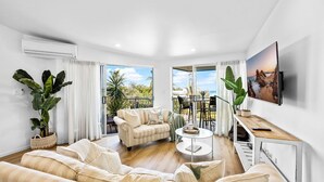 Una sala de estar con sofá, mesa de centro y televisor, con balcón y vistas al exterior.