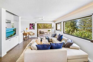 Un salon moderne avec un canapé, une table basse et un téléviseur. Il y a également un coin repas avec des chaises et une table. De grandes fenêtres offrent une vue sur la verdure environnante.
