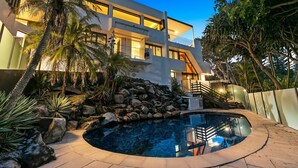 Una casa moderna de varios pisos con piscina, palmeras y un muro de piedra.