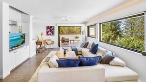 Una sala de estar moderna con sofá, mesa de centro y televisor. Hay una zona de comedor con sillas y una mesa. Grandes ventanas ofrecen vistas al follaje exterior.