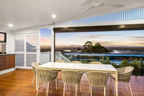 Comedor con mesa y sillas, ventilador de techo y vista a un paisaje a través de amplios ventanales.