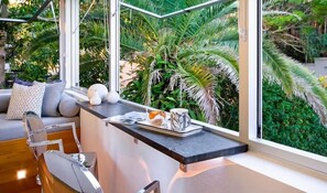 A modern kitchen with a view of lush greenery, a seating area with a sofa and chairs, and a dining table with a tray and mug.