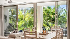 A modern dining area with a wooden table and chairs, large windows, and a view of tropical foliage. - Beautiful Apt in Luxe Beachside Noosa Resort (Noosa)