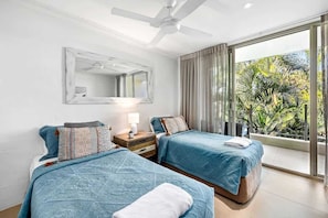 A hotel room with two beds, a ceiling fan, a large window with curtains, and a balcony with a view of greenery.