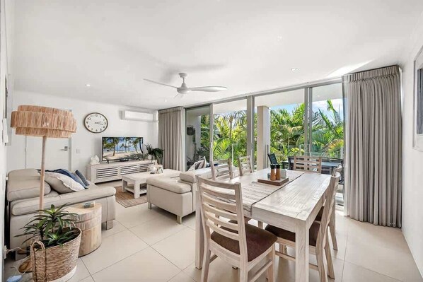 A modern living room with a dining area, large windows, and a ceiling fan.