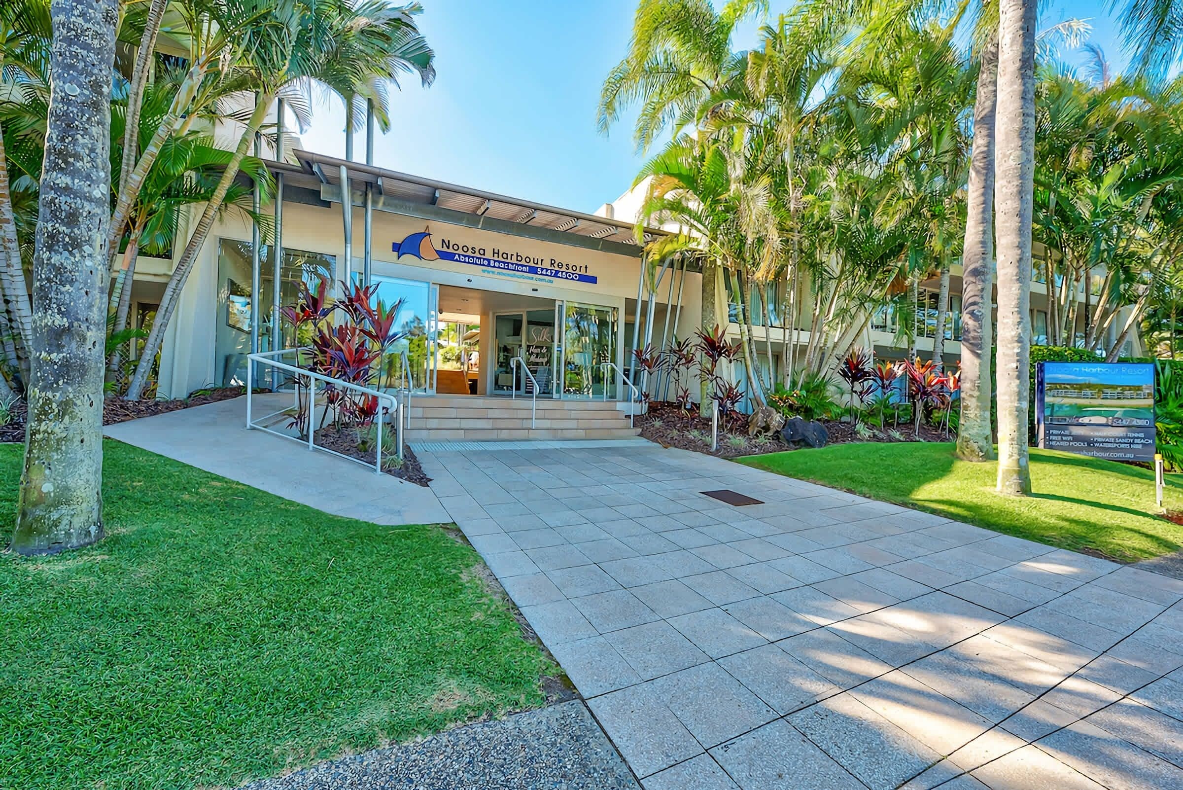 A resort entrance with a sign for Noosa Harbour Resort, featuring a paved walkway, palm trees, and a well-maintained lawn.