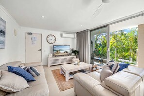 En moderne stue med et fladskærms-tv, beige sofaer, et sofabord og en stor skydedør af glas, der fører ud til en altan med grønt.