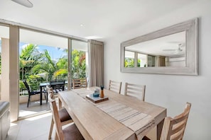A dining area with a wooden table, chairs, and a view of tropical vegetation through large glass doors.