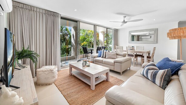 A modern living room with a sofa, coffee table, TV, and a dining area with chairs and a table. - Beautiful Apt in Luxe Beachside Noosa Resort (Noosa)