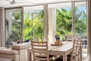 A modern dining area with a glass wall, wooden furniture, and a view of tropical foliage. - Beautiful Apt in Luxe Beachside Noosa Resort (Noosa)