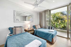 A hotel room with two beds, a ceiling fan, a large window with curtains, and a view of greenery outside.