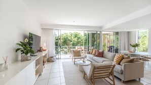 A modern living room with a sofa, coffee table, and chairs, a TV, and a balcony with outdoor furniture. - Heart of Sunshine Spacious Apt Pool and Garden (Noosa)