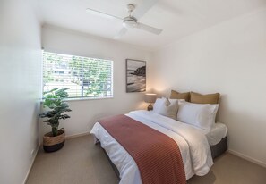 A bedroom with a bed, a ceiling fan, a window with blinds, a painting, and a potted plant.