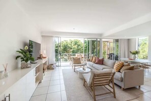 A modern living room with a sofa, coffee table, and TV, a balcony with outdoor furniture, and large windows. - Heart of Sunshine Spacious Apt Pool and Garden (Noosa)