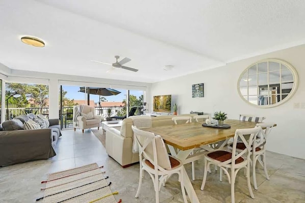 A spacious living area with a wooden dining table, white chairs, a sofa, and a ceiling fan.